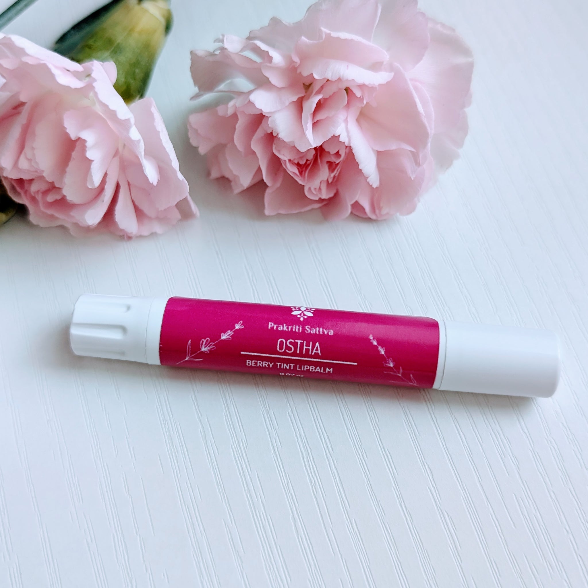 Prakriti Sattva Natural Berry Tinted Lip Balm. A tube of lip balm with a natural berry tint. The label features Ayurvedic symbols and herbs, including alkanet root, indicating the natural ingredients used. There are pink carnation flowers in the background.