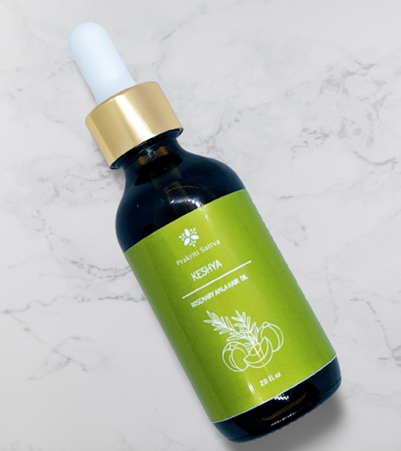 A dark amber bottle filled with Prakriti Sattva Rosemary Amla Hair Oil. The label features Ayurvedic symbols and herbs, including rosemary, amla, bhringaraj, and licorice, indicating the natural ingredients used.