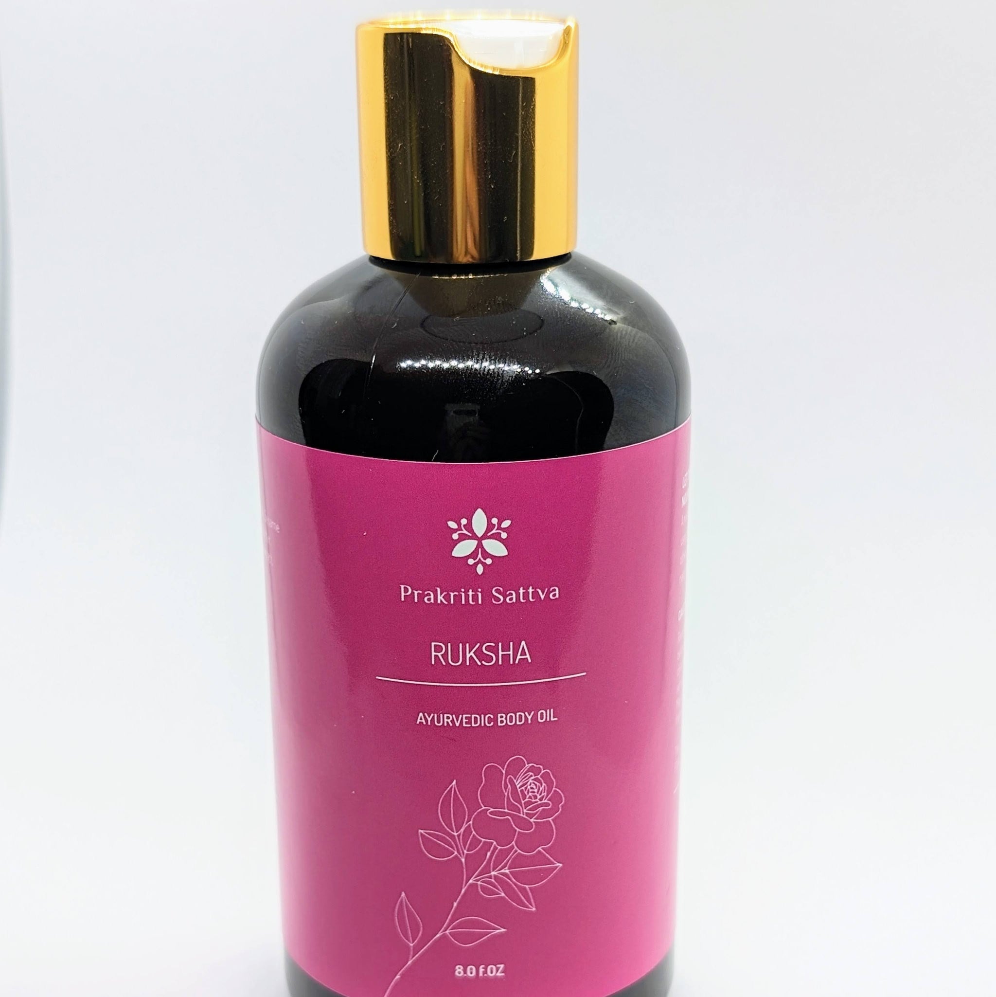A glass bottle filled with Prakriti Sattva Ruksha Ayurvedic Body Oil for Dry Skin. The label features the brand name "Prakriti Sattva" and mentions "Dry Skin." It also features Ayurvedic herbs, including alkanet extract and lavender essential oil, indicating the natural ingredients used.