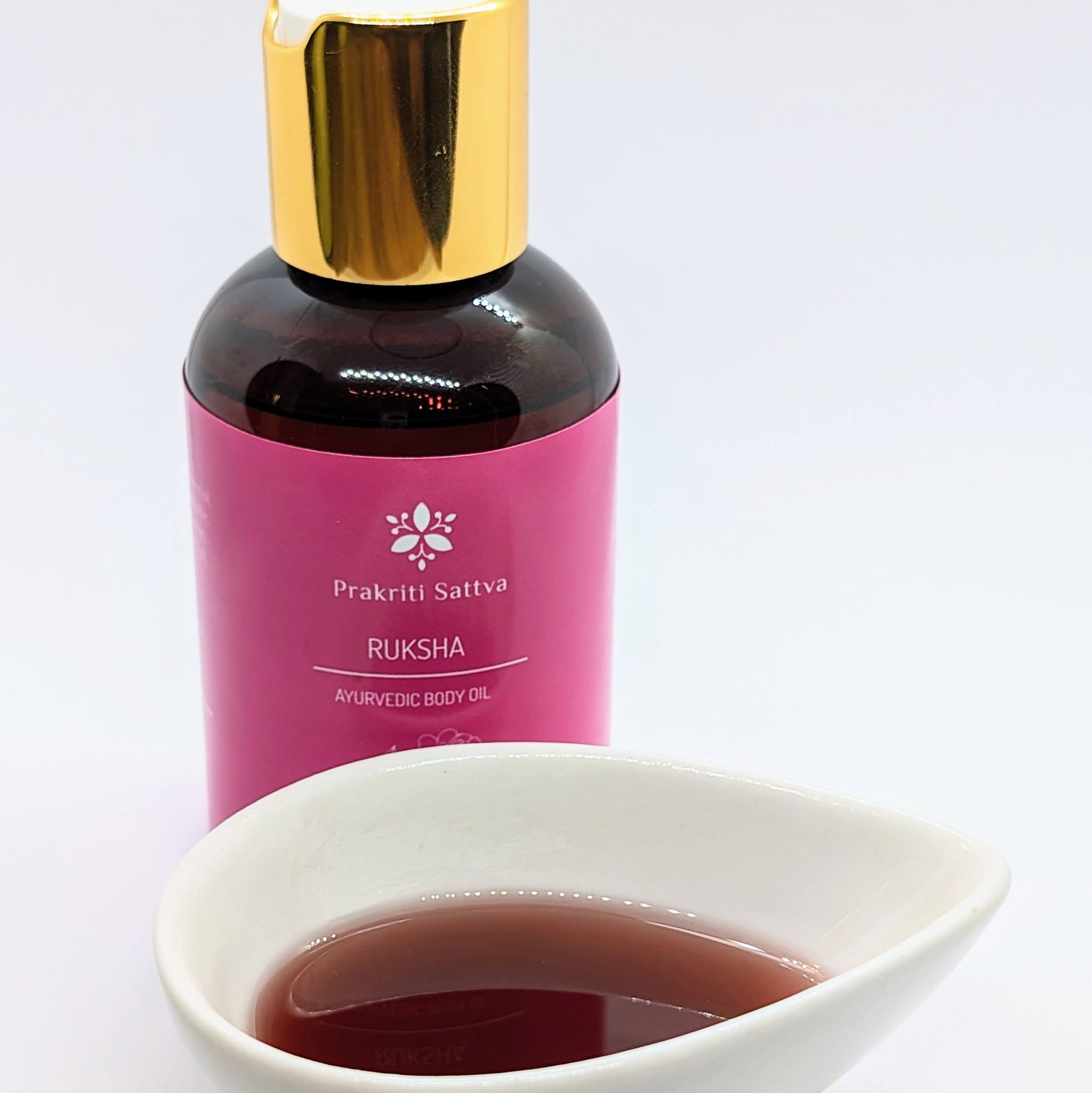 A glass bottle filled with Prakriti Sattva Ruksha Ayurvedic Body Oil for Dry Skin. The label features the brand name "Prakriti Sattva" and mentions "Dry Skin." It also features Ayurvedic herbs, including alkanet extract and lavender essential oil, indicating the natural ingredients used.