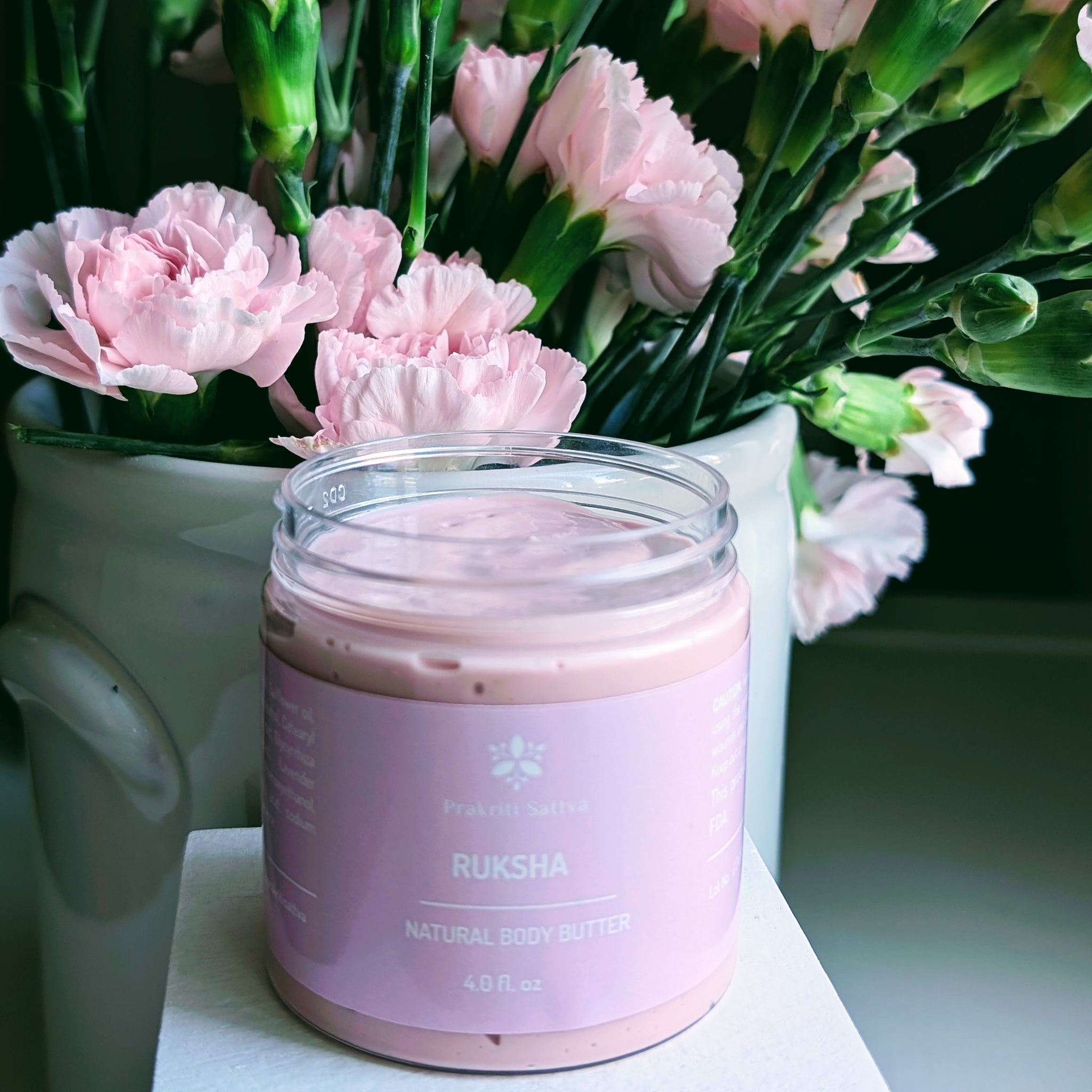 Prakriti Sattva four ounce pink body butter in a plastic jar without the lid. The jar is filled with a creamy, textured, pink body butter product. The background contains pink carnation flowers in a white ceramic vase.