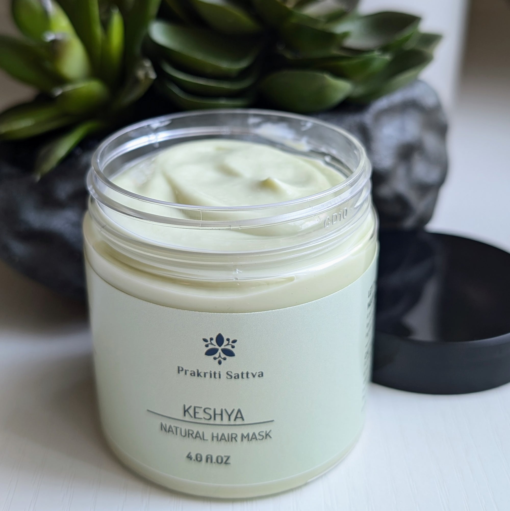 A four ounce jar of Prakriti Sattva Natural Deep Conditioning Hair Mask. The jar is filled with a creamy, green product. The black lid is on the side and there is a succulent plant on the side.