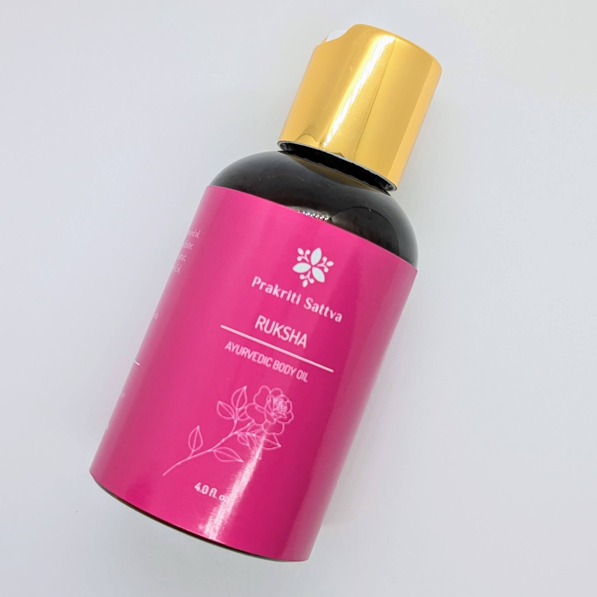A glass bottle filled with Prakriti Sattva Ruksha Ayurvedic Body Oil for Dry Skin. The label features the brand name "Prakriti Sattva" and mentions "Dry Skin." It also features Ayurvedic herbs, including alkanet extract and lavender essential oil, indicating the natural ingredients used.