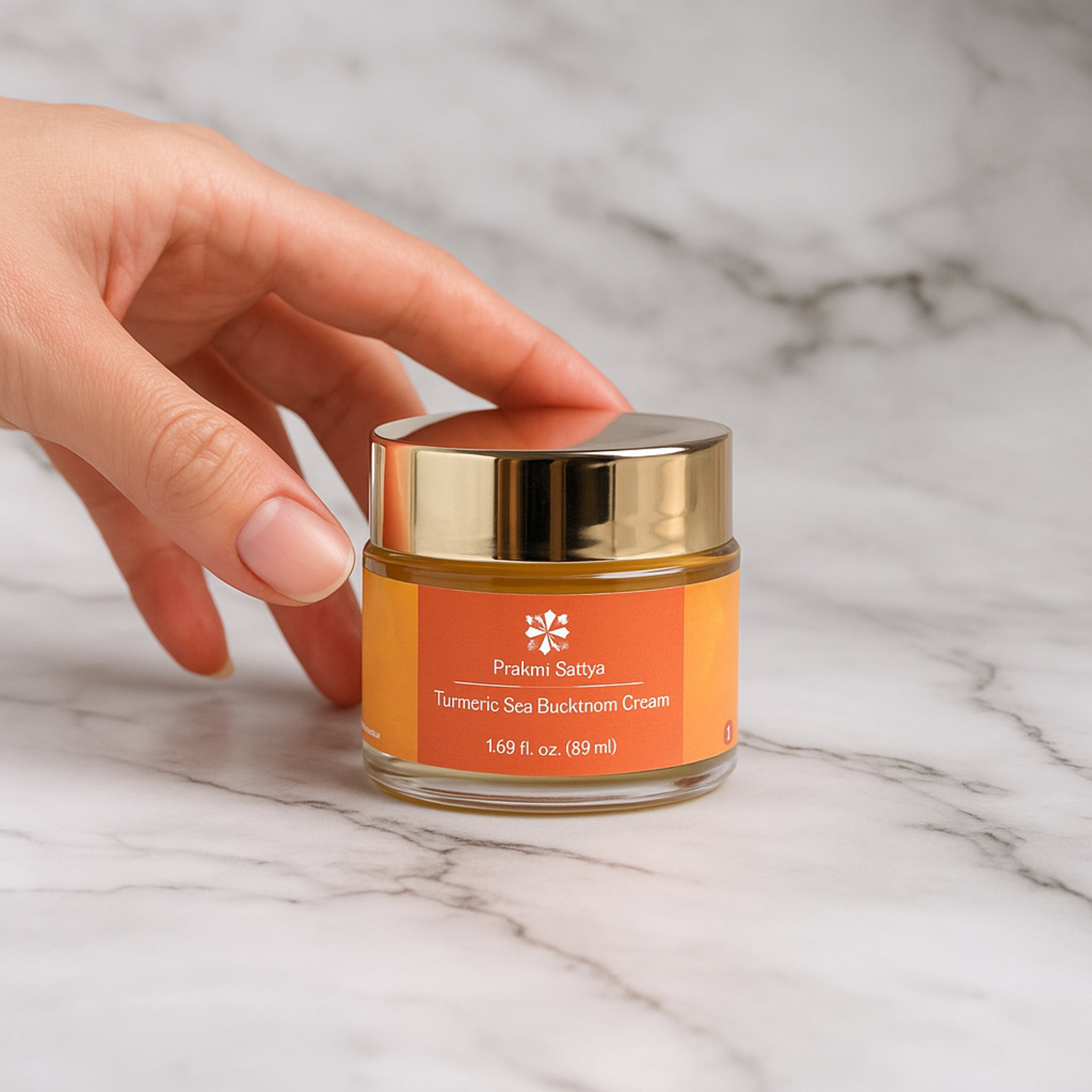 Jar of Prakri Sattva Turmeric Sea buckthorn face cream moisturizer on a marble surface with a hand reaching towards it.
