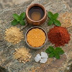 Prakriti Sattva Paduka Intensive repair hand and foot balm in a tin container with a copper bowl containing water and camphor crystals , Indian madder root powder, Indian Sarsaparilla root powder pile on the side on a rock.