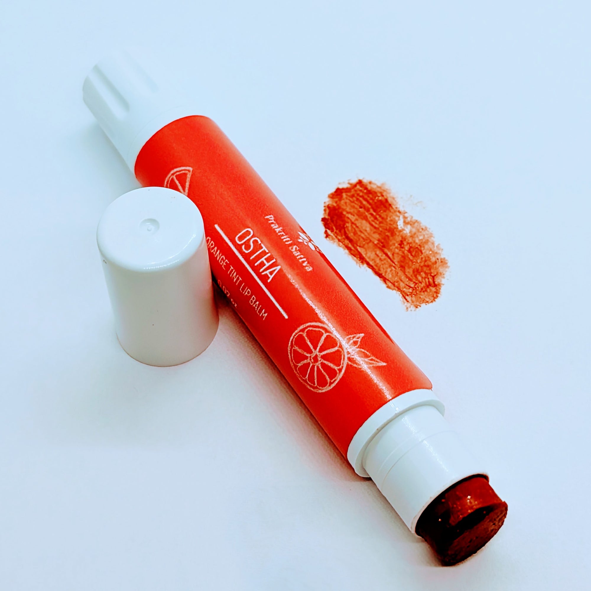 Prakriti Sattva Natural Berry Tinted Lip Balm. A tube of lip balm with a natural berry tint. The label features Ayurvedic symbols and herbs, including Manjistha root, orange, and patchouli, indicating the natural ingredients used. It has a swipe of the lip balm on the side.