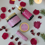 Prakriti Sattva Ruksha body butter, Body oil, Rose face pack and lip balm products with pink labels on a white surface with rose petals and hawthorn berries.