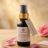 A bottle of Prakriti Sattra Shanti Rose Serum with a gold collar and clear pump top, styled on a wooden surface with pink rose petals and a pink rose in the background.