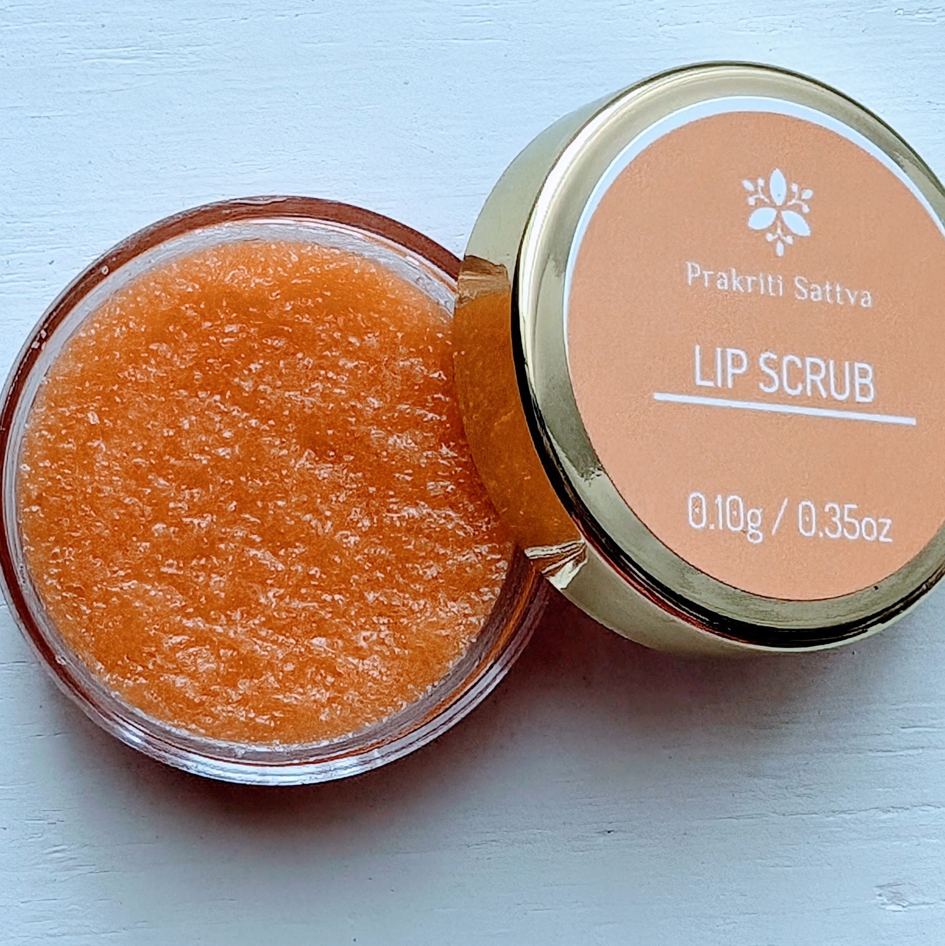 A small container of Prakriti Sattva Ayurvedic Natural Sugar lip scrub in orange color with lid open.