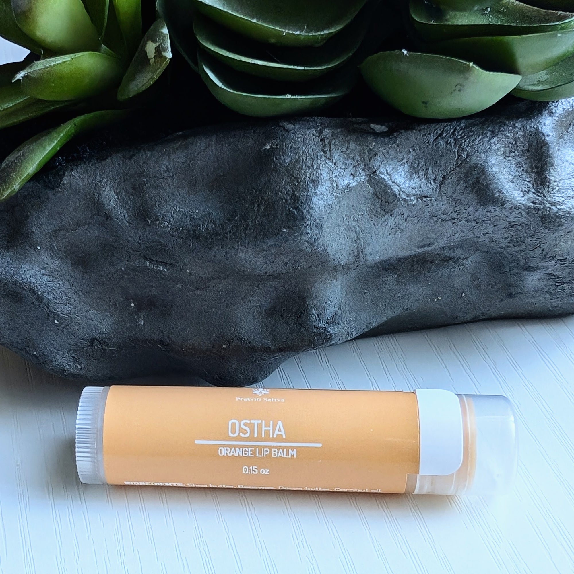 Ayurvedic natural lip balm with Manjistha (Indian madder root) extract, orange essential oil, and patchouli essential oil. It is a tube of lip balm with a natural, earthy color. The label features Ayurvedic symbols and herbs, including Manjistha, orange, and other essential oils, indicating the natural ingredients used.