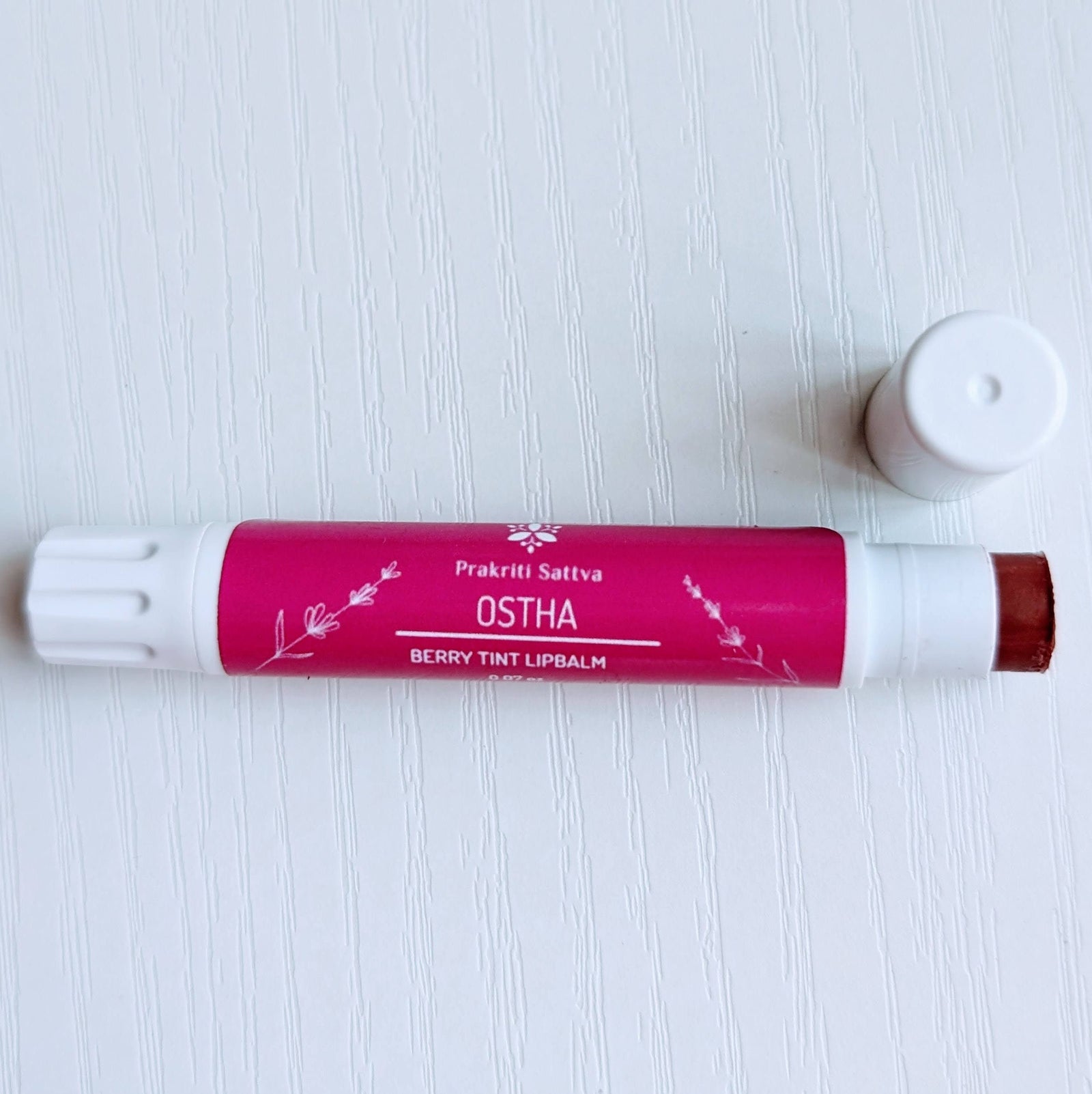 Prakriti Sattva Natural Berry Tinted Lip Balm. A tube of lip balm with a natural berry tint. The label features Ayurvedic symbols and herbs, including alkanet root, indicating the natural ingredients used.