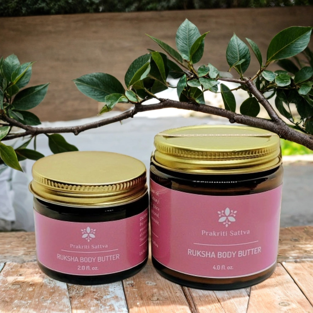 Two ounce and four ounce jars of Prakriti Sattva Ruksha Body Butter on a wooden surface with green leaves in the background.