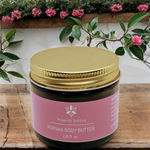 Two ounce amber glass Jar of Ruksha Body Butter with pink label and gold lid on a wooden surface with plants in the background