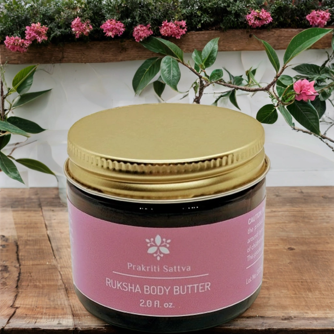 Two ounce amber glass Jar of Ruksha Body Butter with pink label and gold lid on a wooden surface with plants in the background