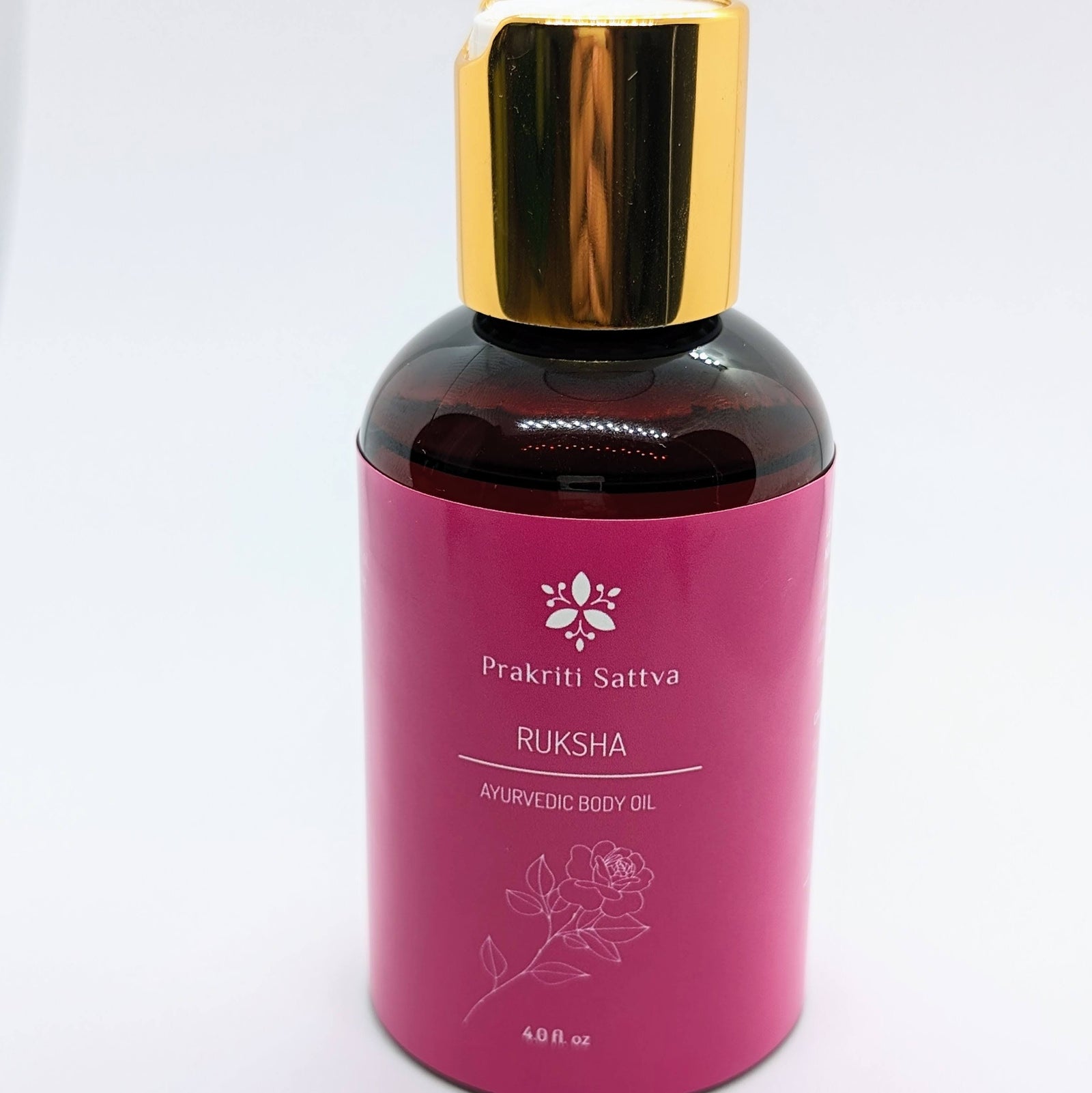 A glass bottle filled with Prakriti Sattva Ruksha Ayurvedic Body Oil for Dry Skin. The label features the brand name "Prakriti Sattva" and mentions "Dry Skin." It also features Ayurvedic herbs, including alkanet extract and lavender essential oil, indicating the natural ingredients used.