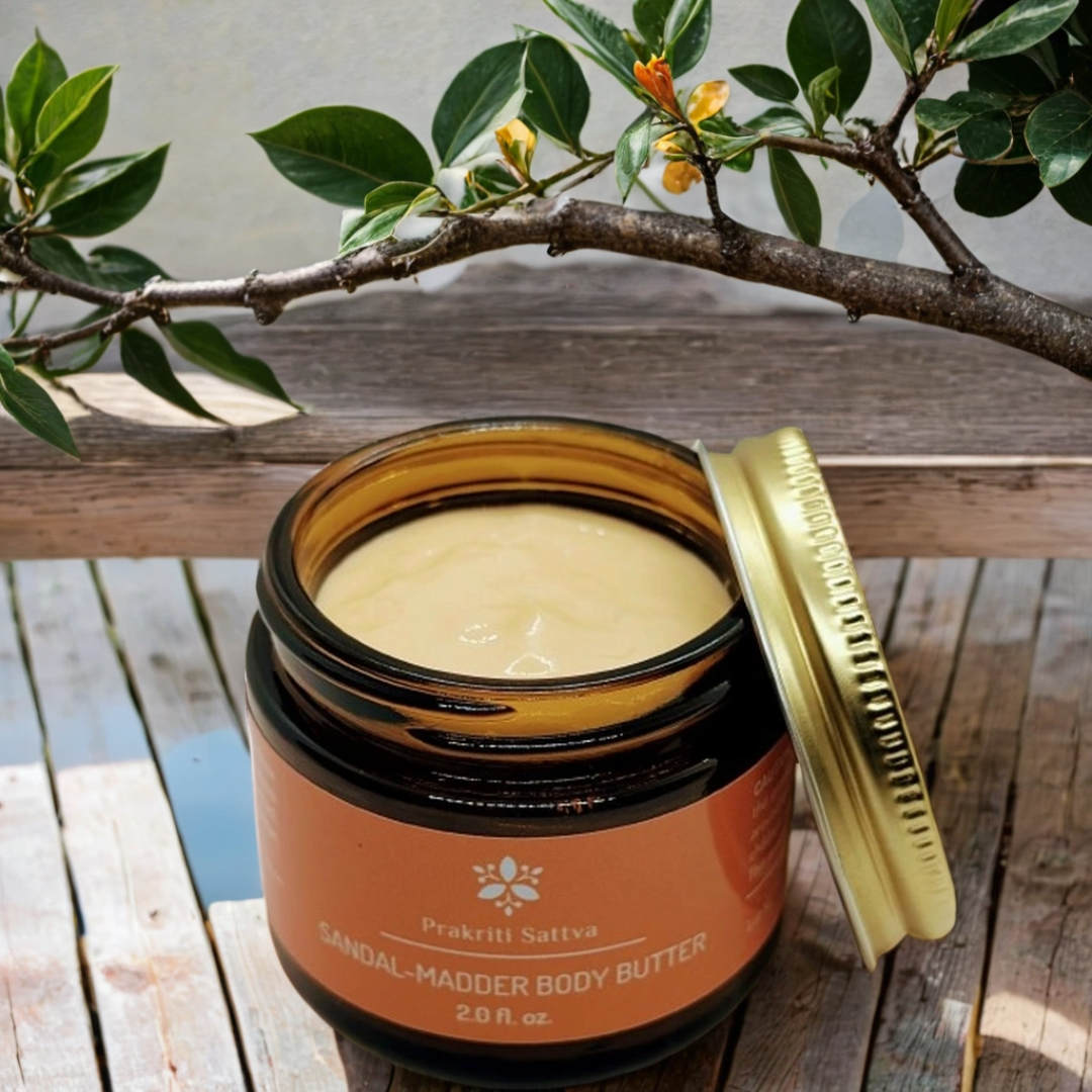 Jar of Sandalwood Madder Body Butter with lid open and lying on the side on a wooden surface with green leaves in the background