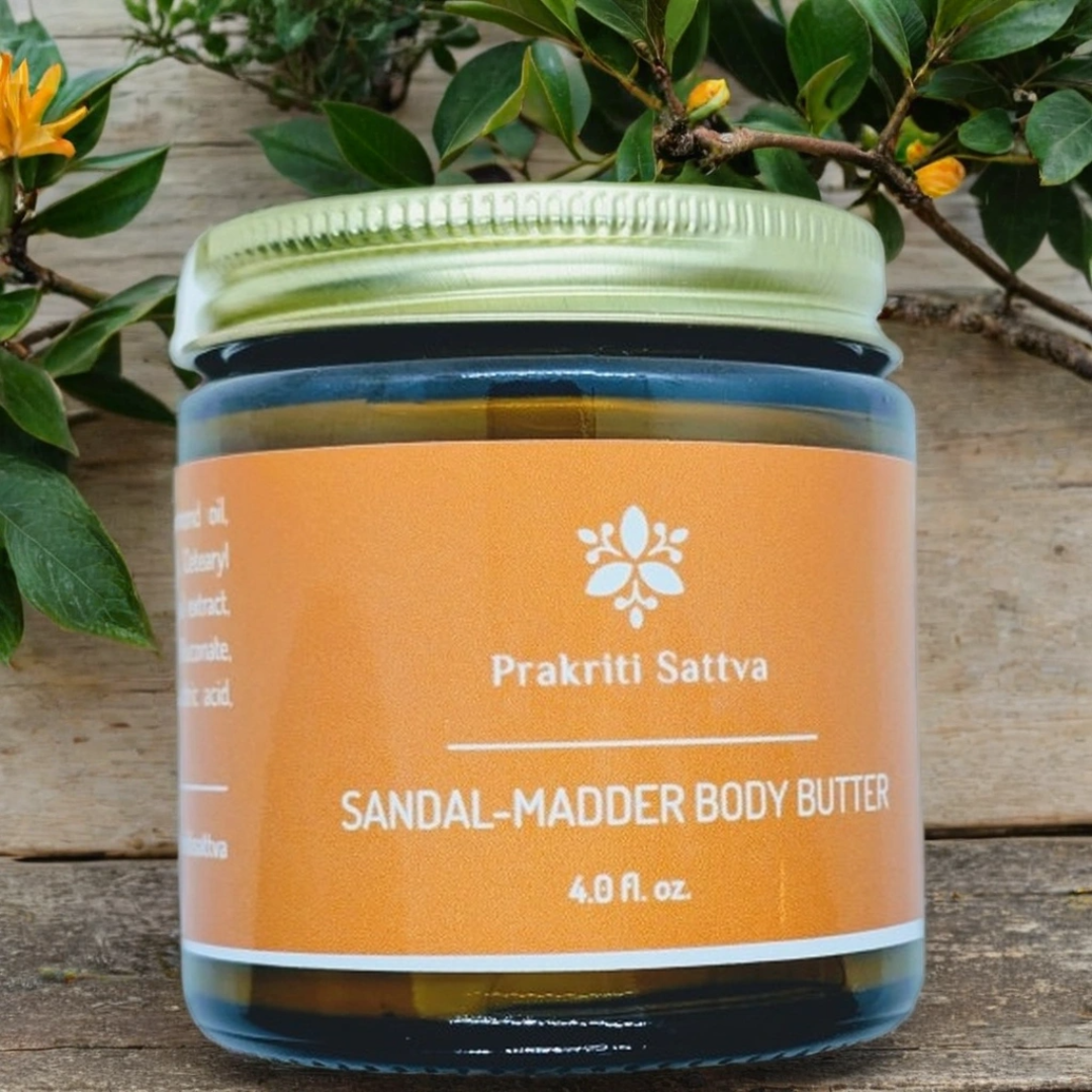 Four ounce Jar of Prakriti Sattva Sandal-Madder Body Butter on a wooden surface with greenery in the background