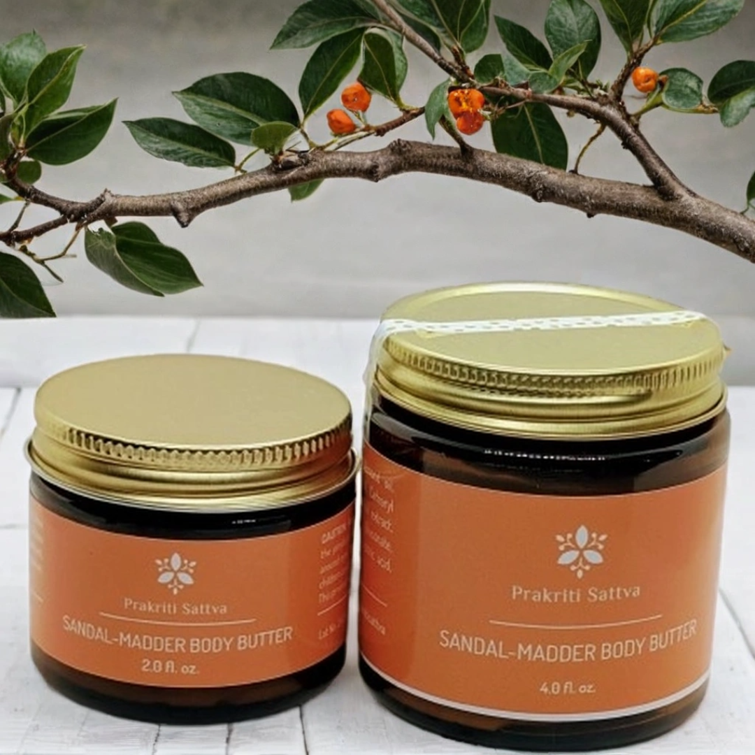 Two jars of Prakriti Sattva Sandal-Madder Body Butter on a white surface with green leaves in the background.