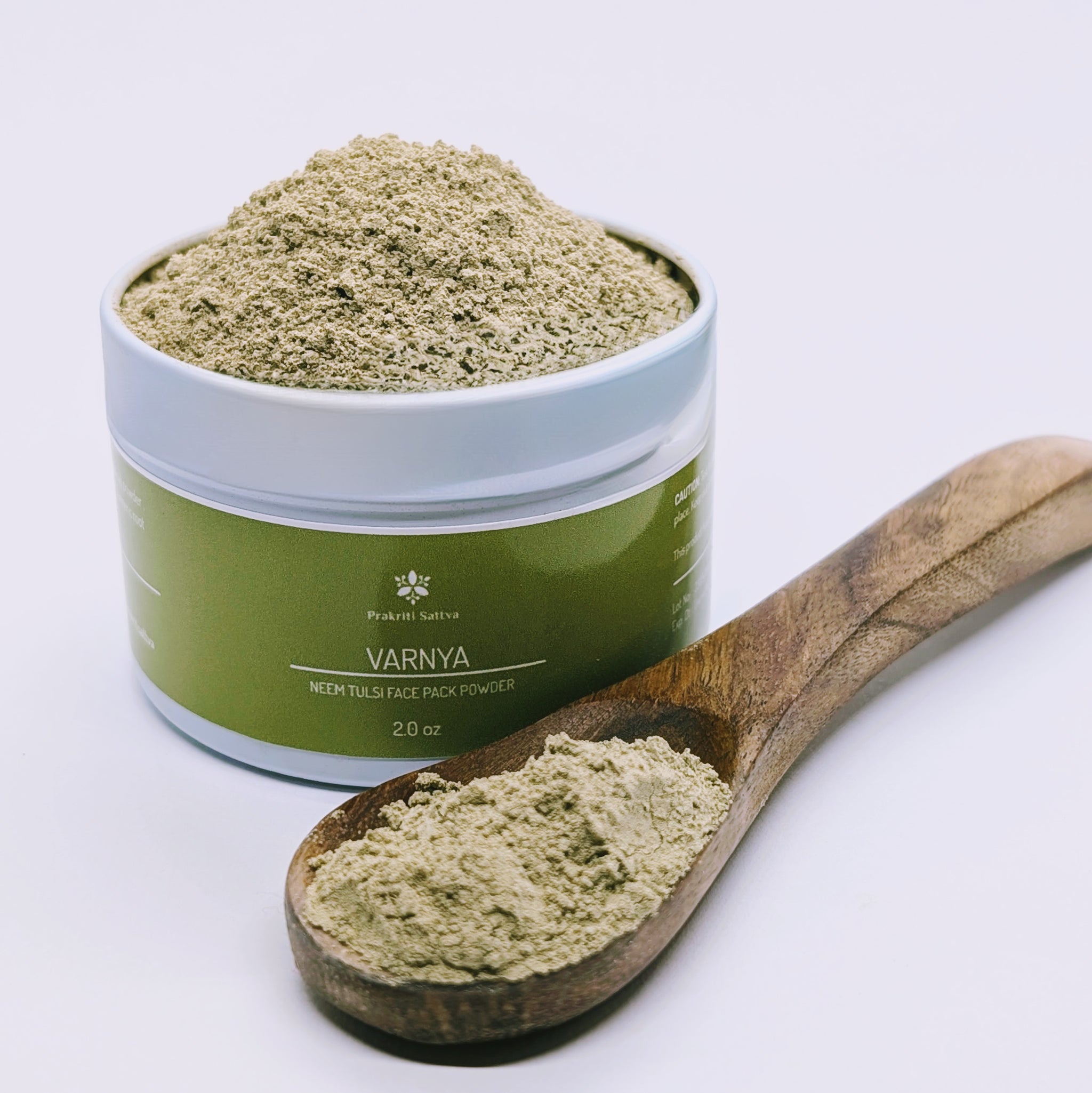 An open 2 ounce aluminum jar of Prakriti Sattva Neem Tulsi Moringa Amla Face Pack for Acne-Prone and Kapha-Pitta Skin. A wooden spoon with a scoop of the powder face pack in front. A natural, Ayurvedic face mask to help reduce acne, balance oil production, and soothe inflammation.